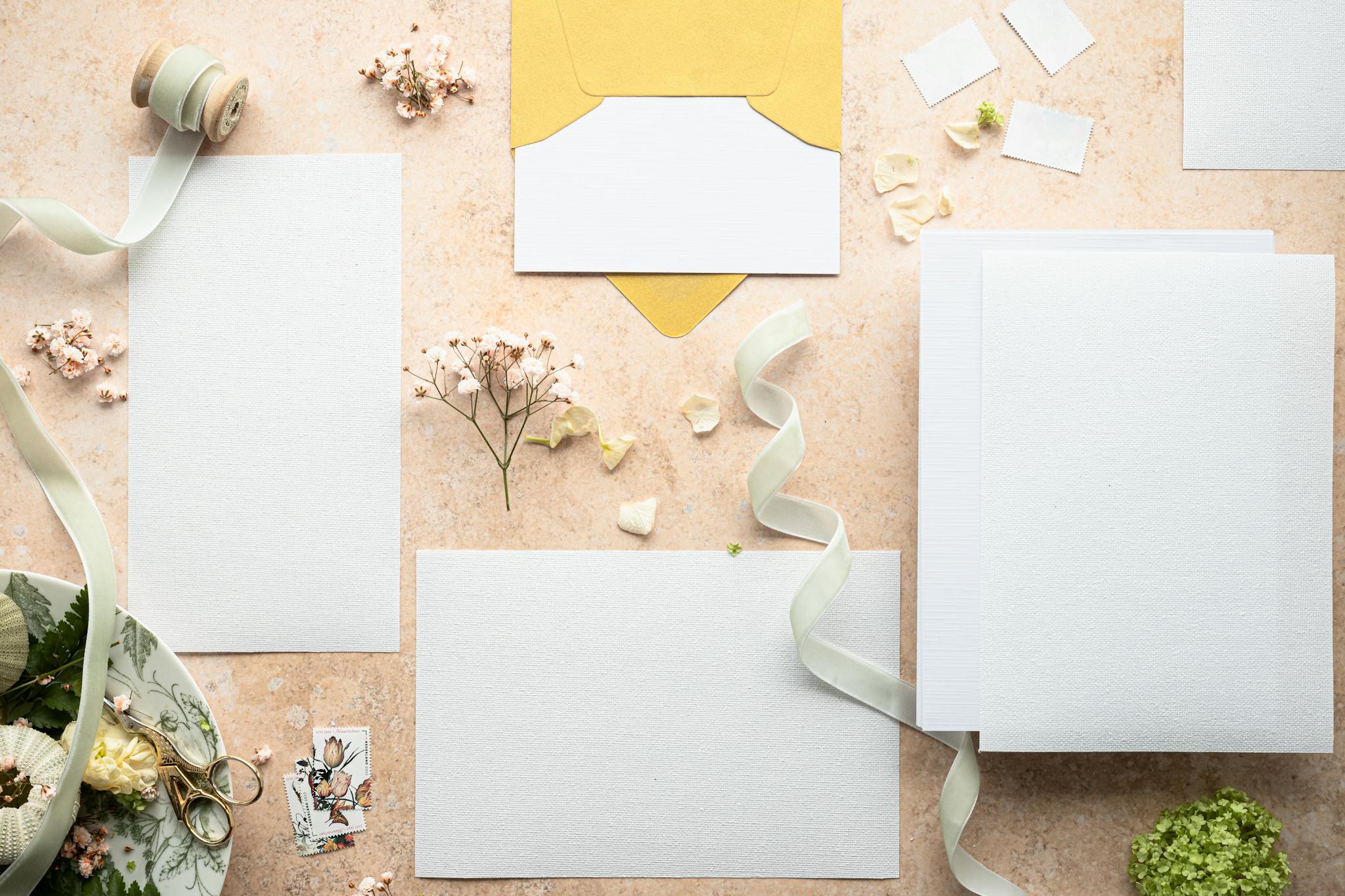 A flat lay of stationery, flowers, ribbons, and envelopes on a textured surface.