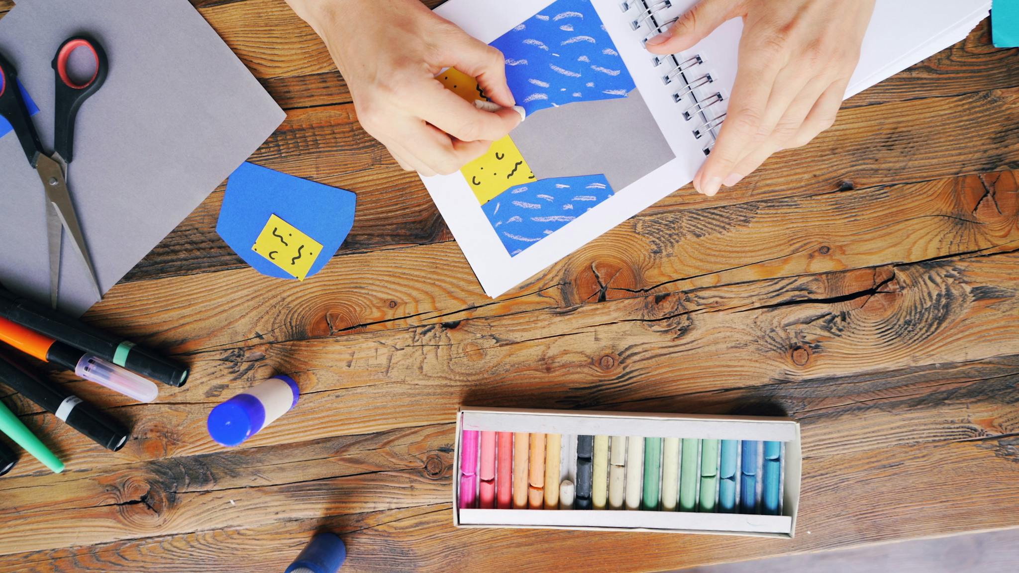 Hands crafting with colored paper and pastels on a wooden table.